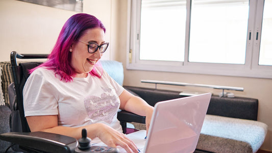 A woman with striking pink hair uses a pink laptop. She is sitting in a power wheelchair