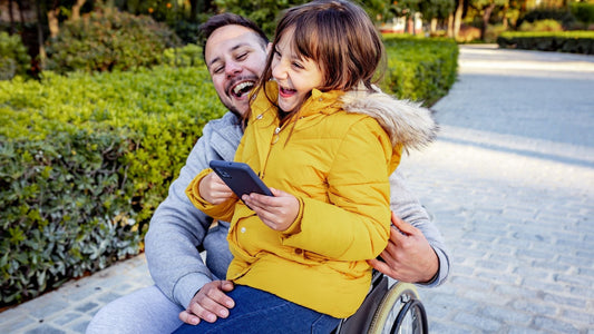 Girl in yellow coat sits on wheelchair user's lap. Both are laughing.
