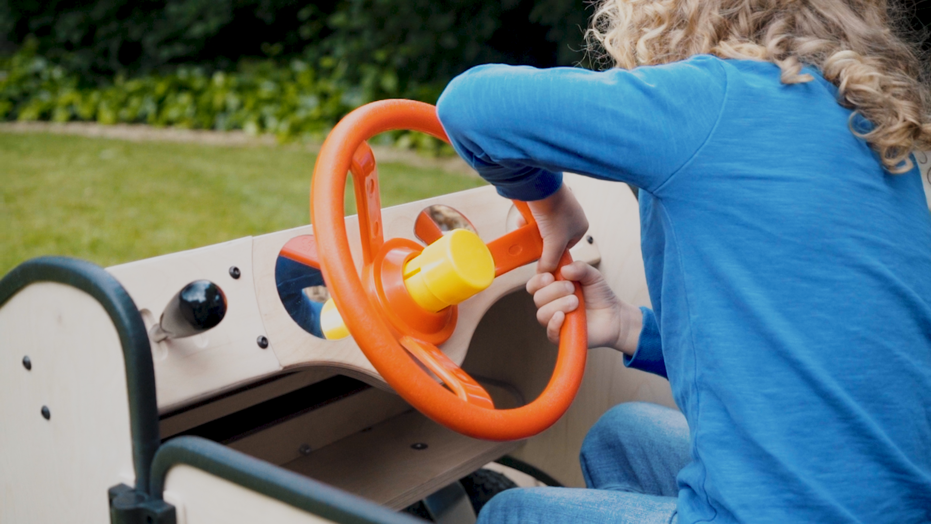 A child wearing a blue top is driving the Big Car. They face away from the camera and lean to the side holding two hands on one side of the stering wheel as if they are steering round a corner.