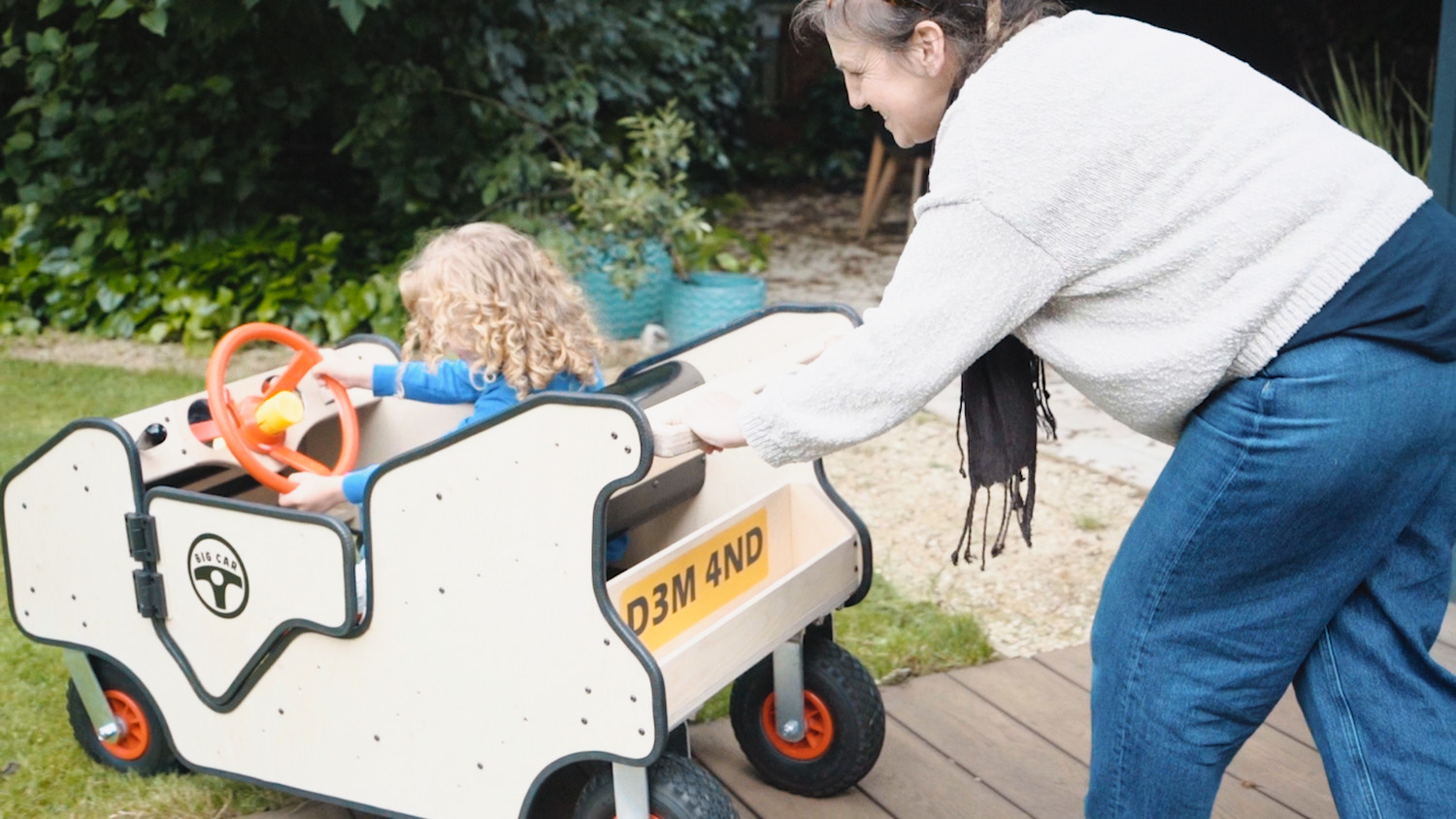 An adult holds the piush bar on the Big Car while a child with curly blonde hair is sat inside. The child holds the steering wheel.
