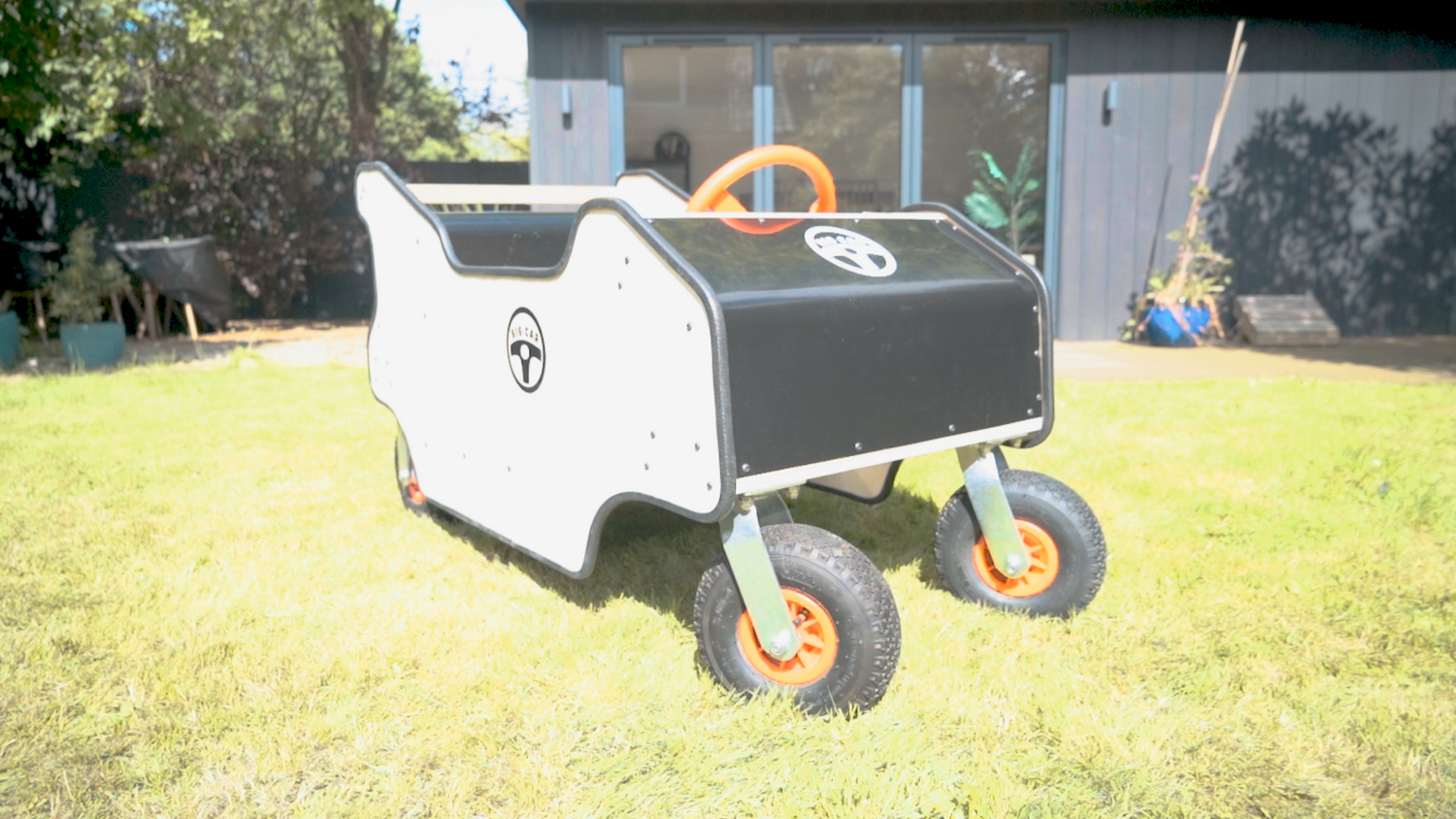 The Big Car is pictured from the front corner showing the entire car. The side is light wood colour and the front is shiny black plastic. A logo saying Big Car is on the front and Side and a red steering wheel is partially visible.