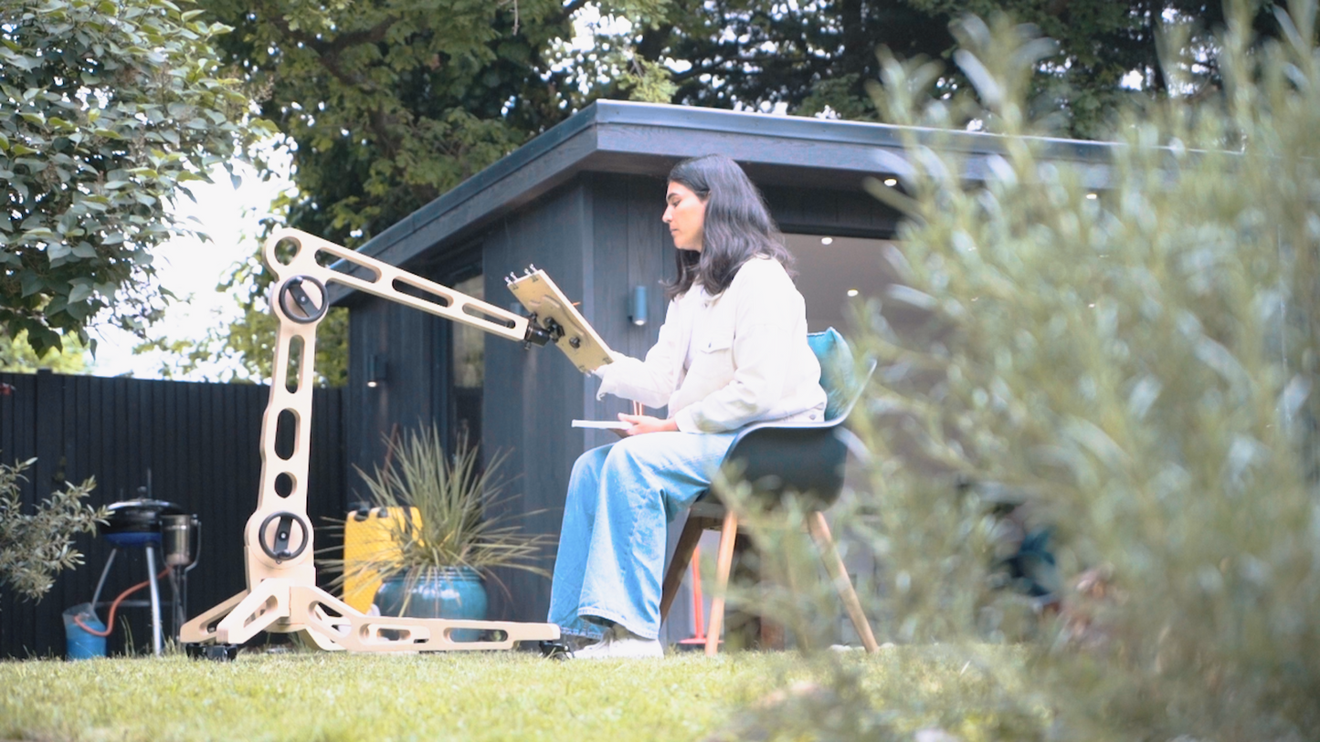 A female sits on a chair in a garden space and paints on a board held by the Freasel. It is positioned to hold the painting surface at a height reached eaily from her seated position.
