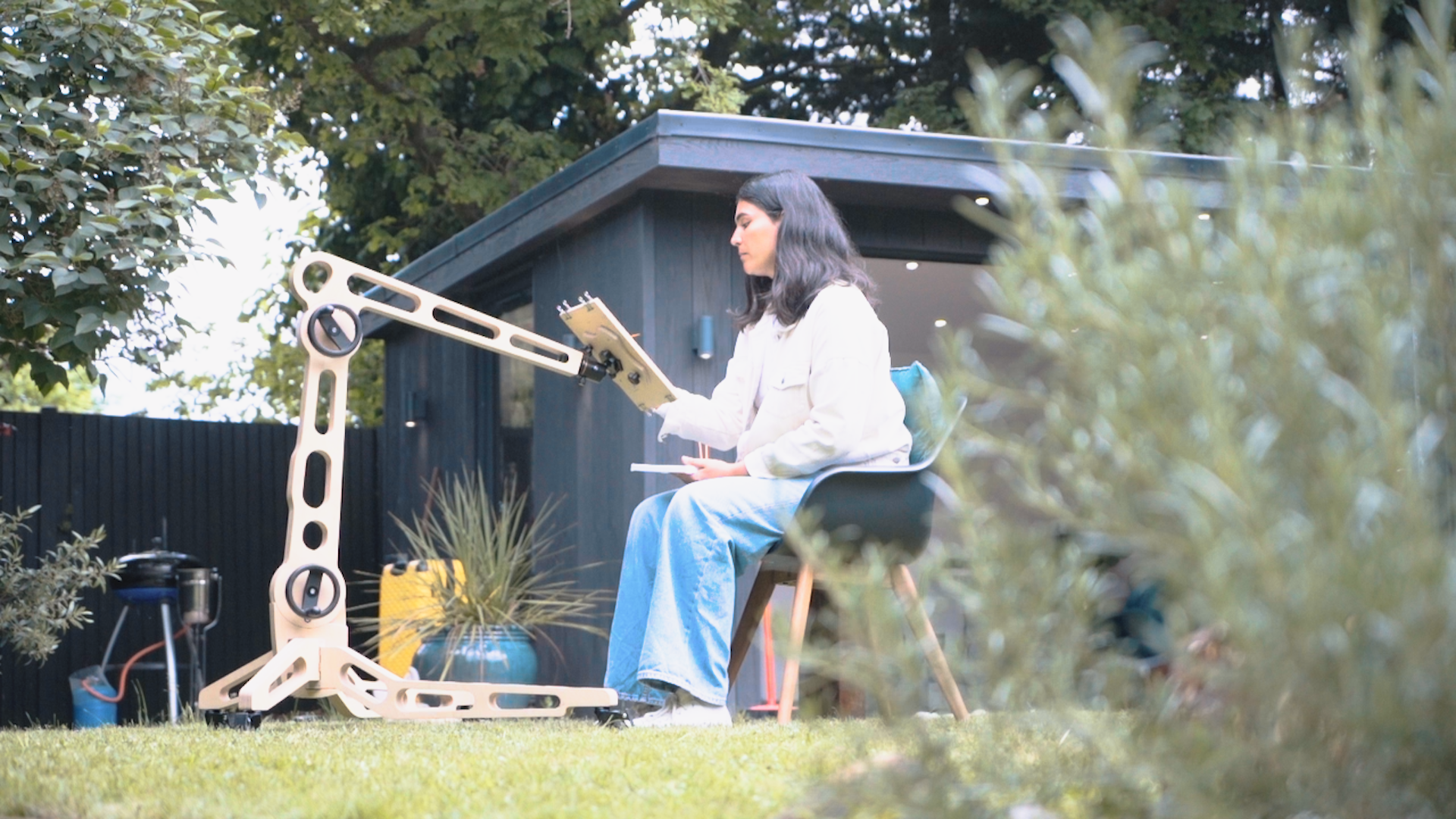 A female sits on a chair in a garden space and paints on a board held by the Freasel. It is positioned to hold the painting surface at a height reached eaily from her seated position.