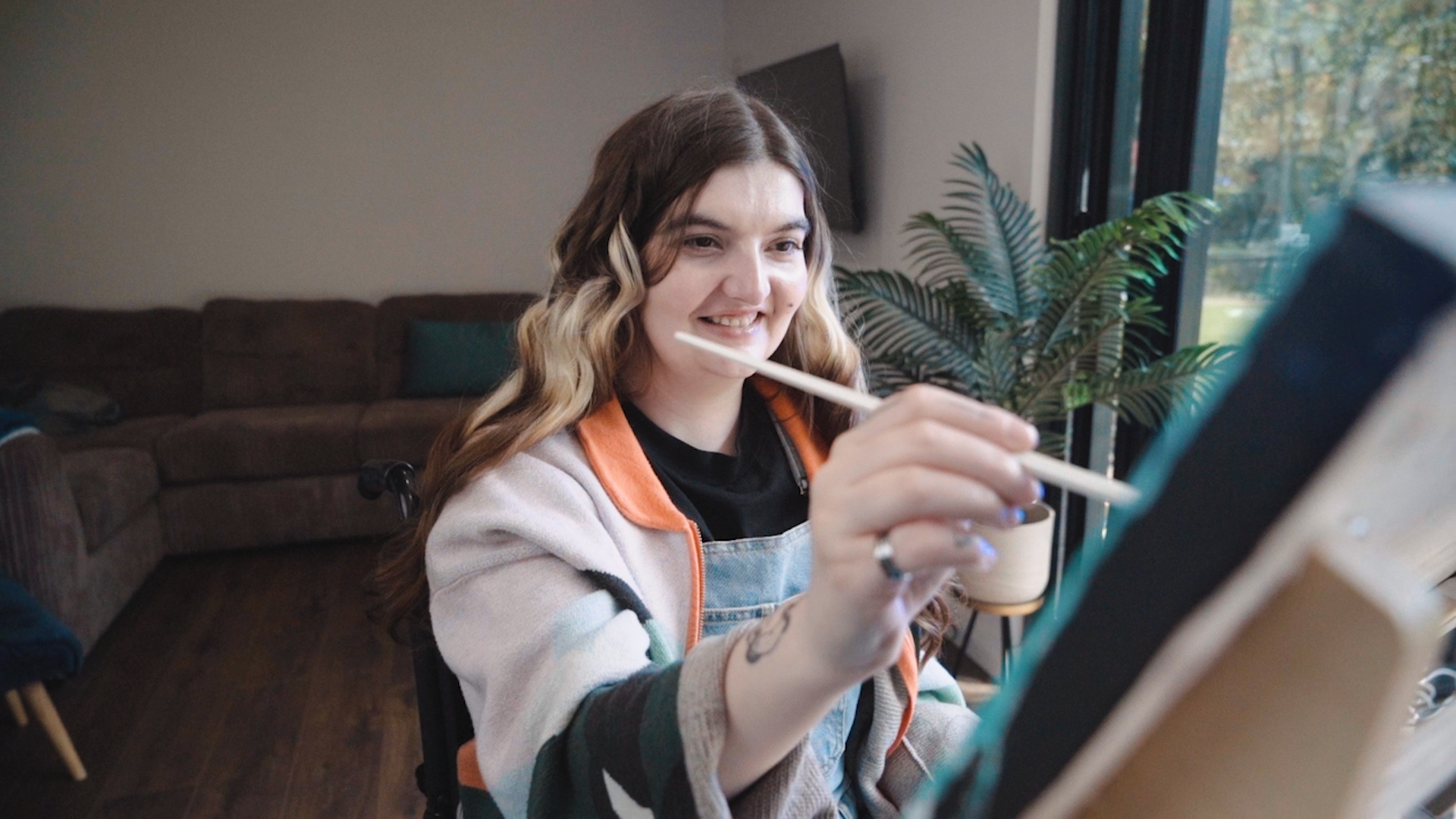 A person with long wavy hair faces the camera while they touch a paintbrush to a canvas held on a freasel which is not in the image.