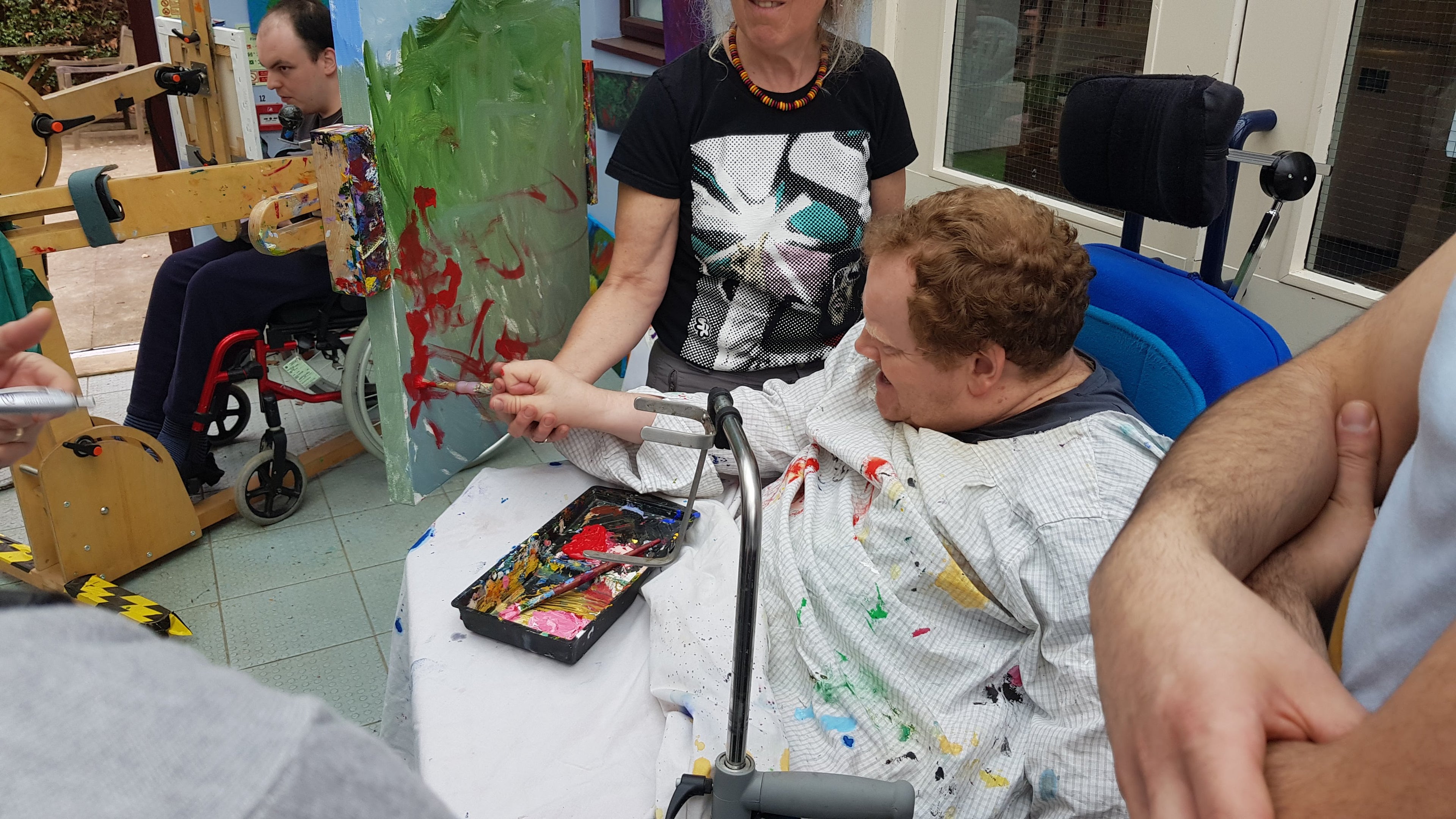 A painter in a wheelchair is side on to a canvas on the Freasel with another person steadying their hand while they apply red paint