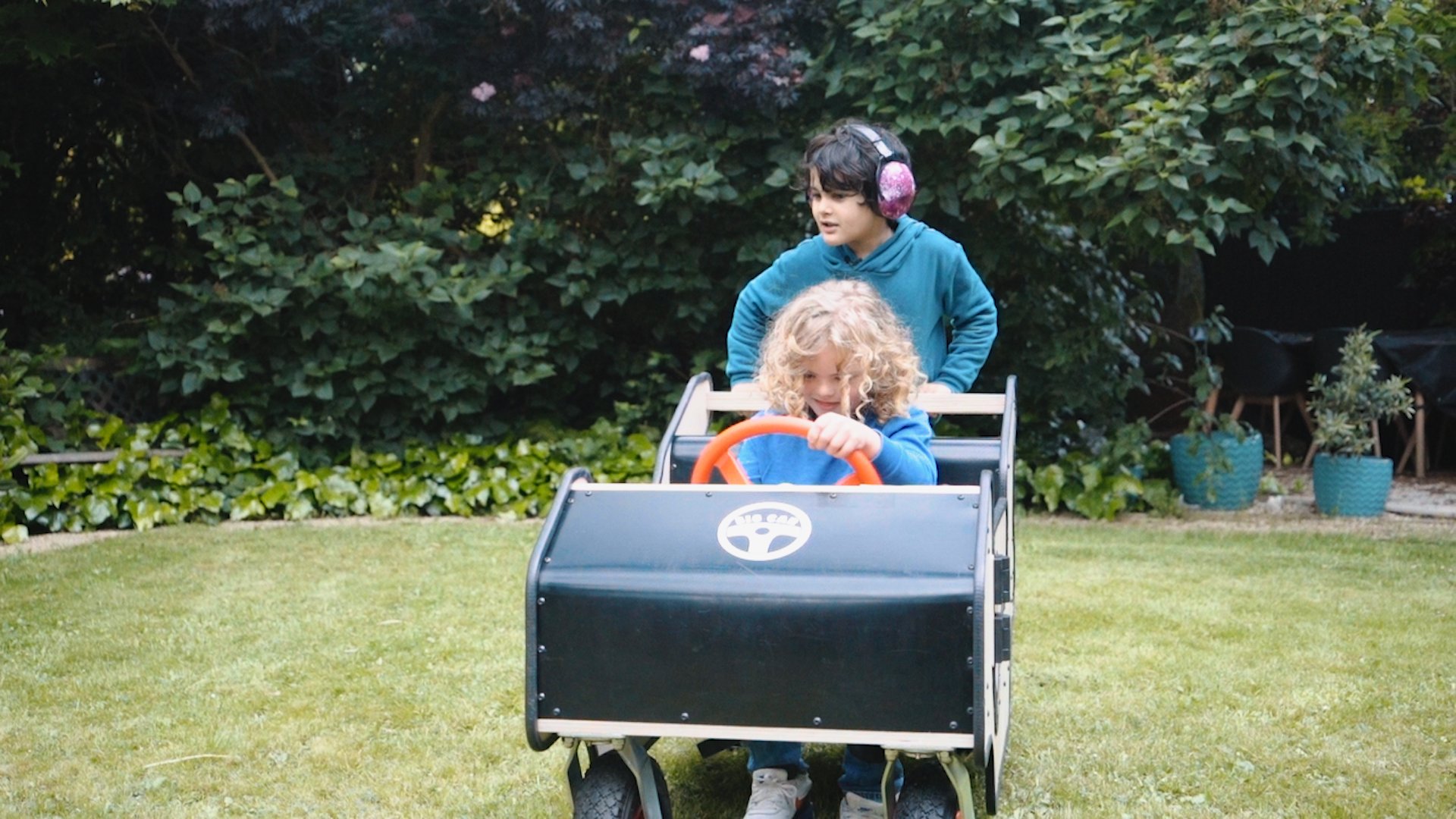 Two children around 6 -8 years old play with the Big Car in a garden. One child with curly blonde hair sits in the car and holds the steering wheel. Another child with short dark hair and purple ear defenders stands at the back fo the car holding the push bar. 
