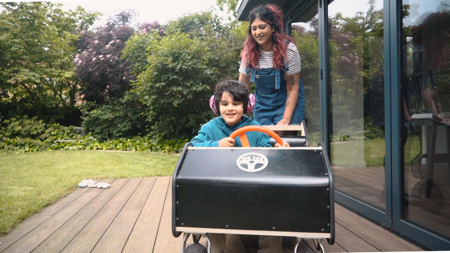 An adult and child play together on a wooden deck with the Big Car. The adult stands behind the child in the car holding the push bar. The smiling child looks ahead at the direction the car is moving. They wear purple ear defenders and a teal blue top. 