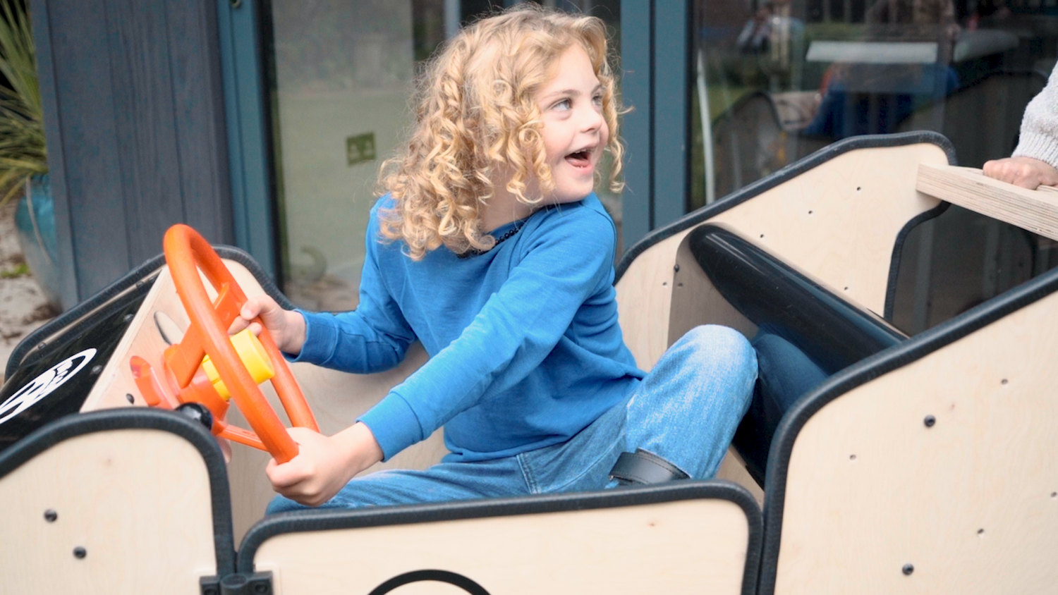 A child is sitting in the big car and looks back over their shoulder at the adult behind them. They grin as they hold the red steering wheel.