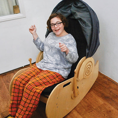A teenager sits in the sensory shell chair with the hood up. Her arms are raised