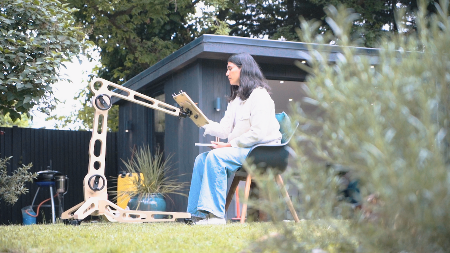 A female sits on a chair in a garden space and paints on a board held by the Freasel. It is positioned to hold the painting surface at a height reached eaily from her seated position. 