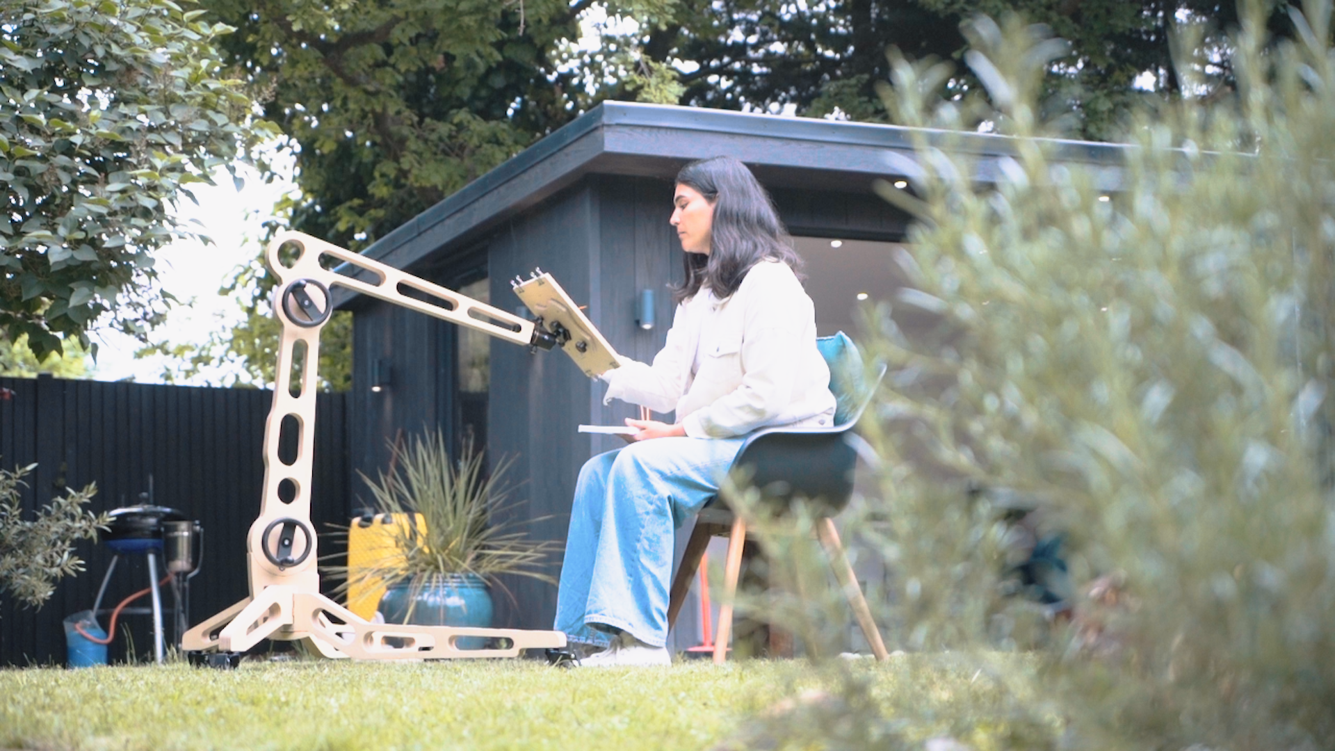 A female sits on a chair in a garden space and paints on a board held by the Freasel. It is positioned to hold the painting surface at a height reached eaily from her seated position. 