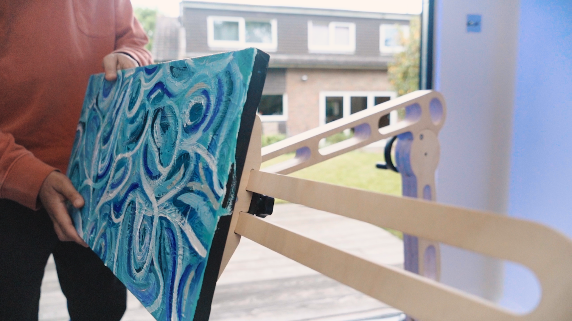 A parsons hands hold a canvas painted with swirls of blue in all shades. The canvas is fixed to a Freasel accessible easel.