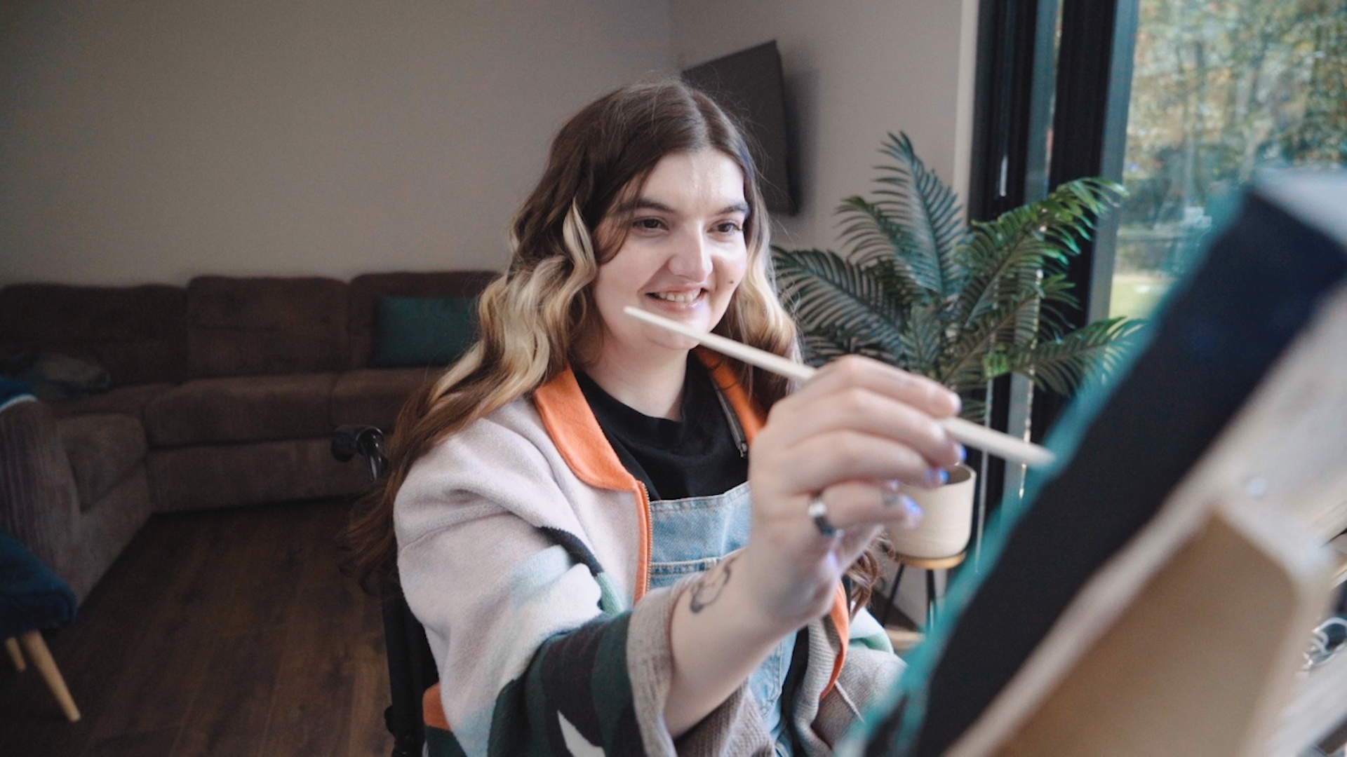 A person with long wavy hair faces the camera while they touch a paintbrush to a canvas held on a freasel which is not in the image. 