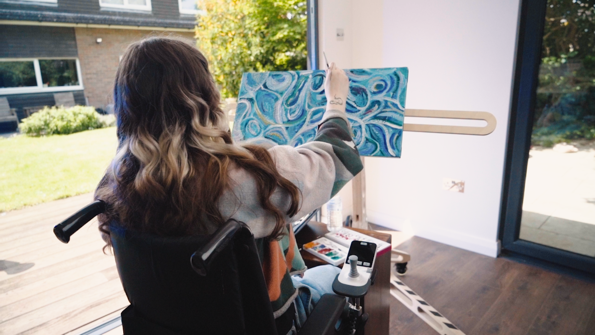 A person is seated in a wheelchair with push handles and a joystick. They face away from the camera while painting on a canvas with vabious shades of blue swirls which is mounted on a freasel. 