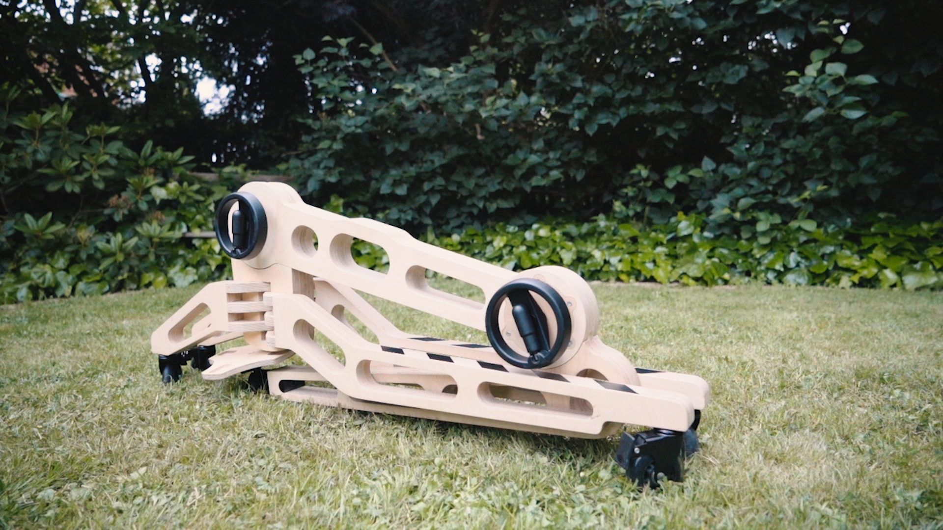 The freasel is outside on some grass in it's folded state. It's a skeletal form made of light coloured plywood. Two circular black adjustment handles are facing the camera.