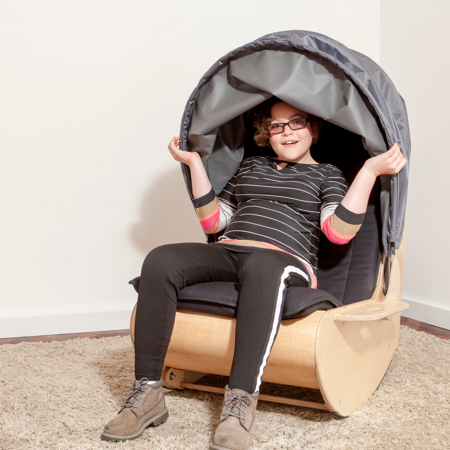 Teenager sits in sensory shell rocking chair with hands on hood to pull it down 