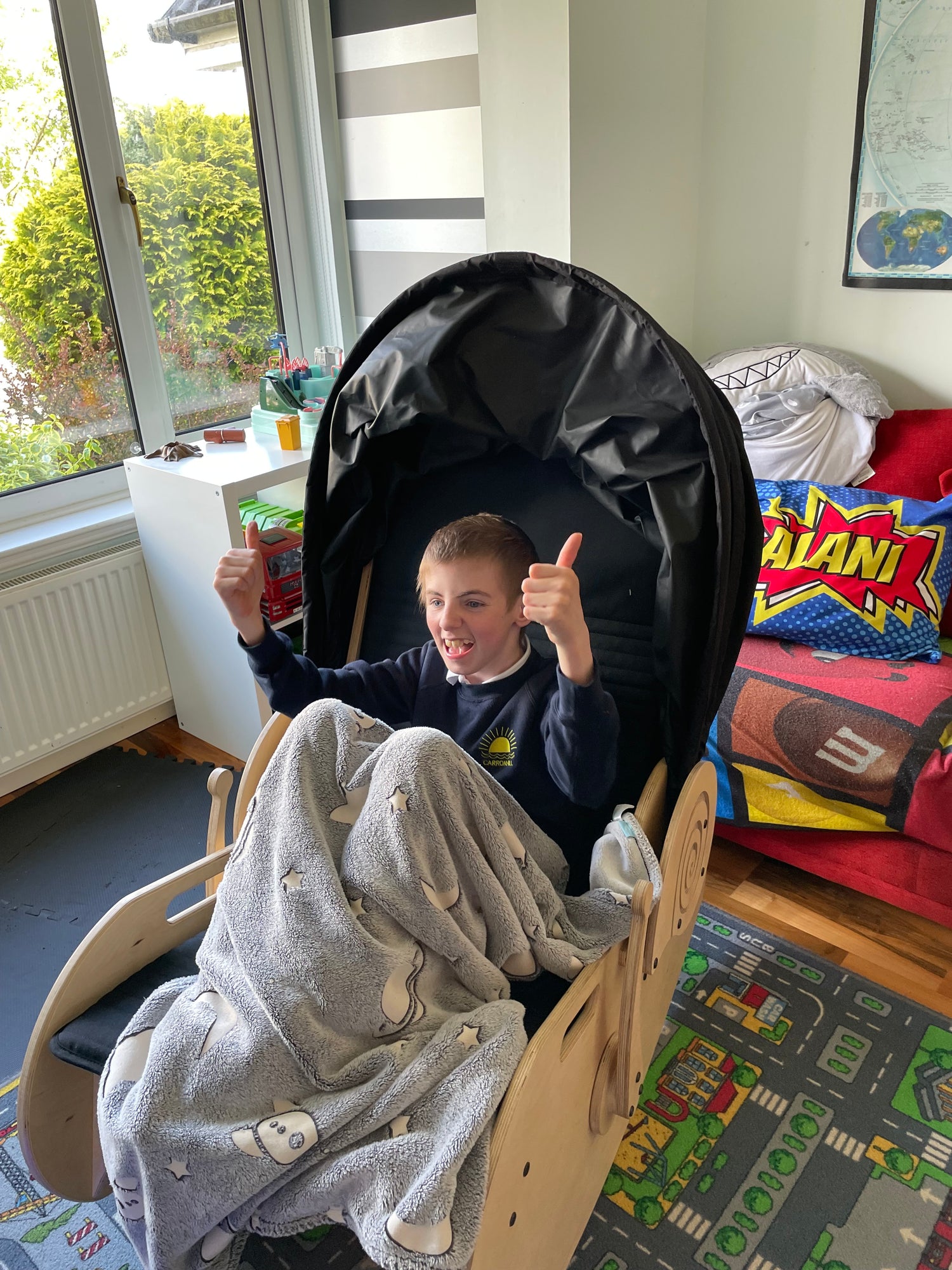 A child sits in the sensory shell chair with a blanket over his knees giving double thumbs up