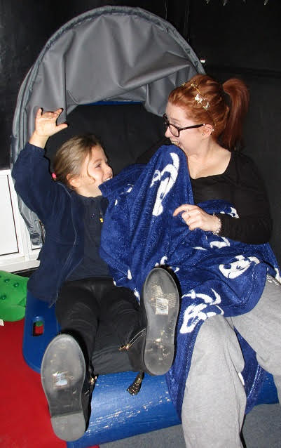 A child sits in the sensory shell chair with their feet up as the chair rocks. An adult sits nest to them with a blanket