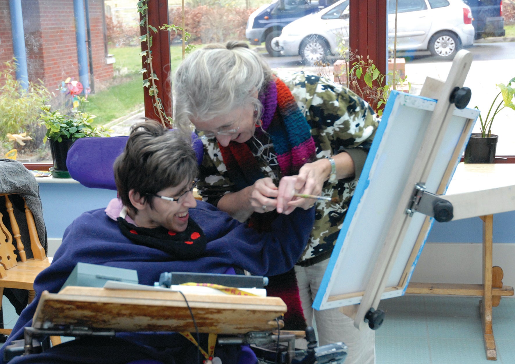 A painter in their wheelchair smiles and is reaching to the canvas with help of another person holding the brush tightly